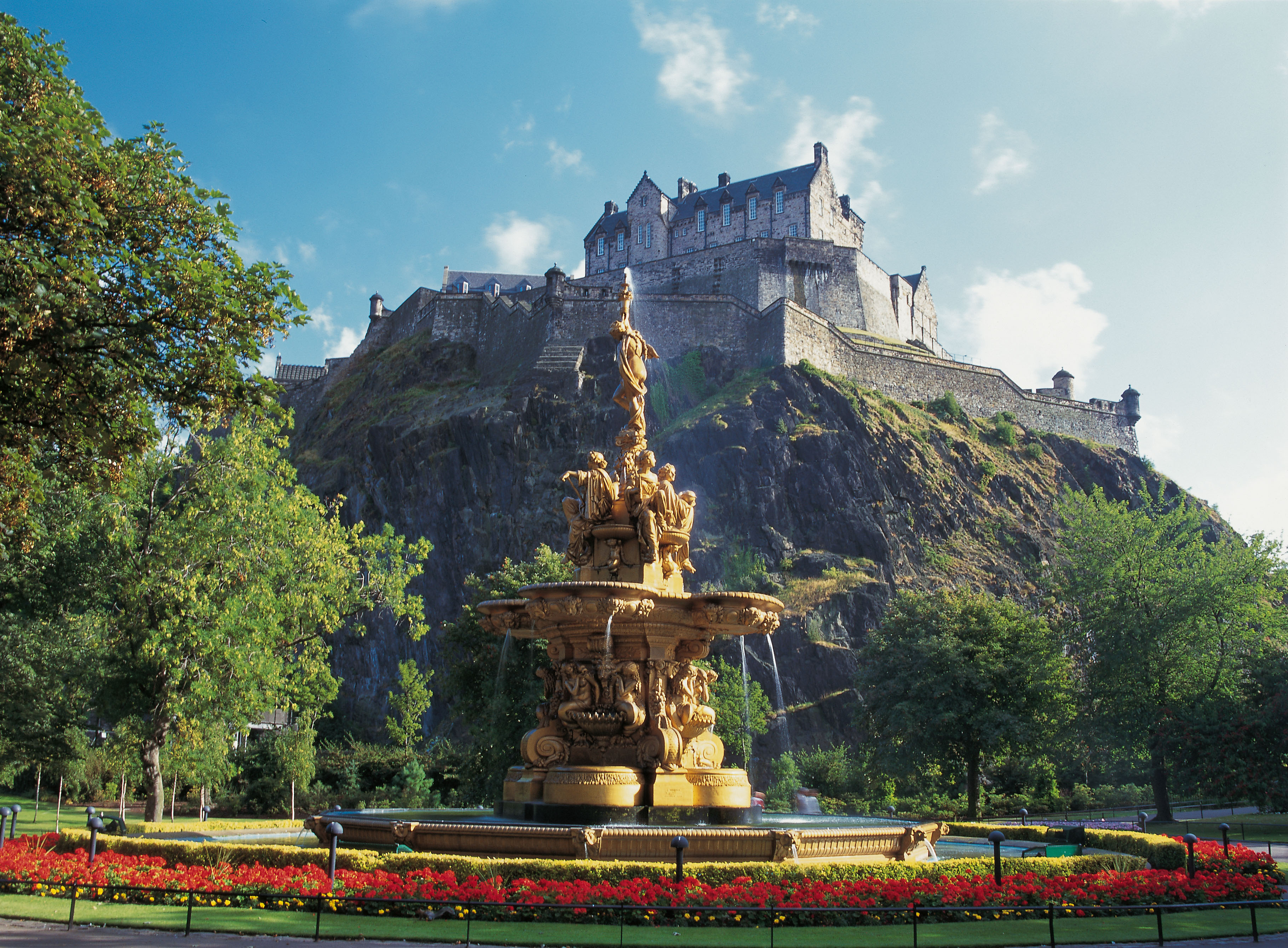 Photograph of Edinburgh Castle taken from Princes Street Gardens with an ornate golden water fountain in the foreground and blue sky and sunshine